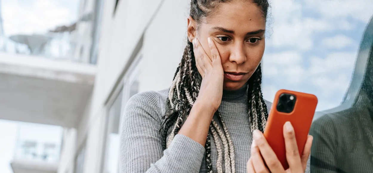 Young woman with long braids, hand on her cheek, looking worried at a smartphone in an orange case